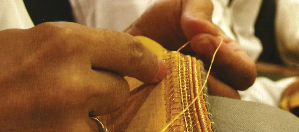 Hands stitching gold zari onto the edge of a bisht